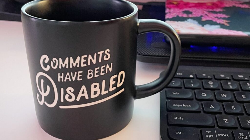 A mug with the text, "Comments Have Been Disabled" on it, next to a keyboard and a laptop on a cooling mat behind it