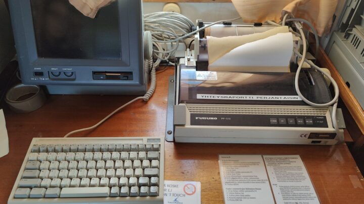 A vintage ship communication setup with a CRT monitor, keyboard, and printer, along with printed instructions, likely for maritime navigation and communication.