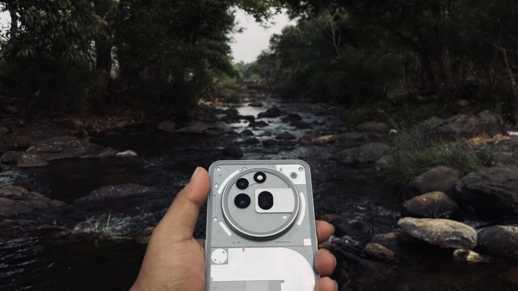 A hand holds a transparent smartphone, showing internal components, against a serene forest backdrop with a flowing stream.