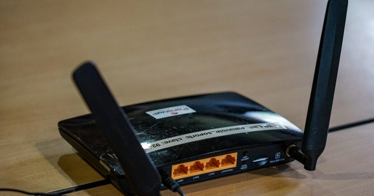 A black wireless router with two antennas on a wooden surface with visible status lights and a cable plugged in.