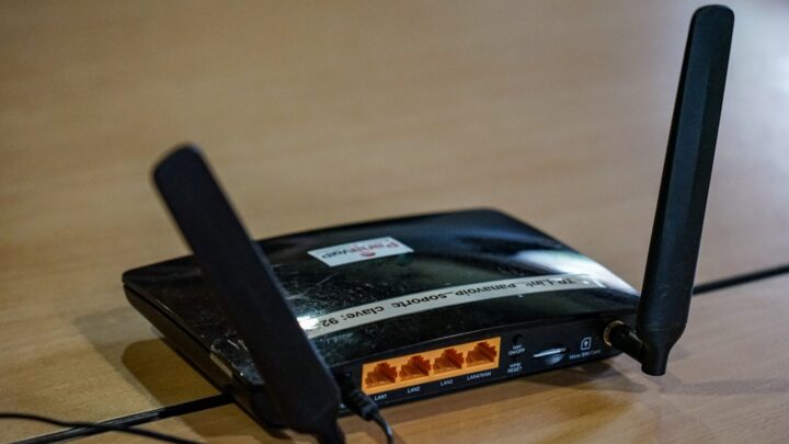 A black wireless router with two antennas on a wooden surface with visible status lights and a cable plugged in.