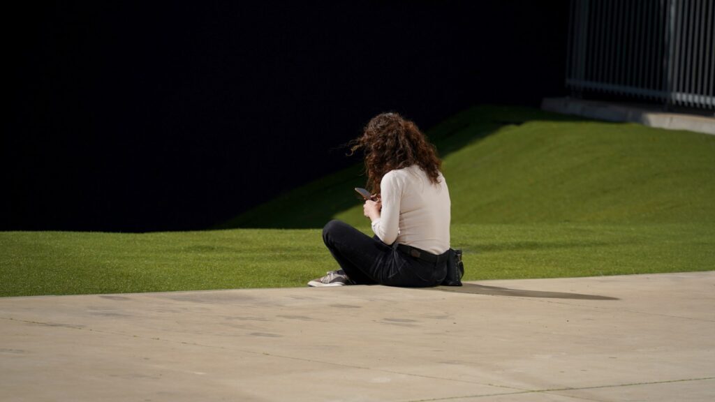 A person with curly hair wearing a white long-sleeve shirt and black pants is sitting on a grassy area, using a smartphone.