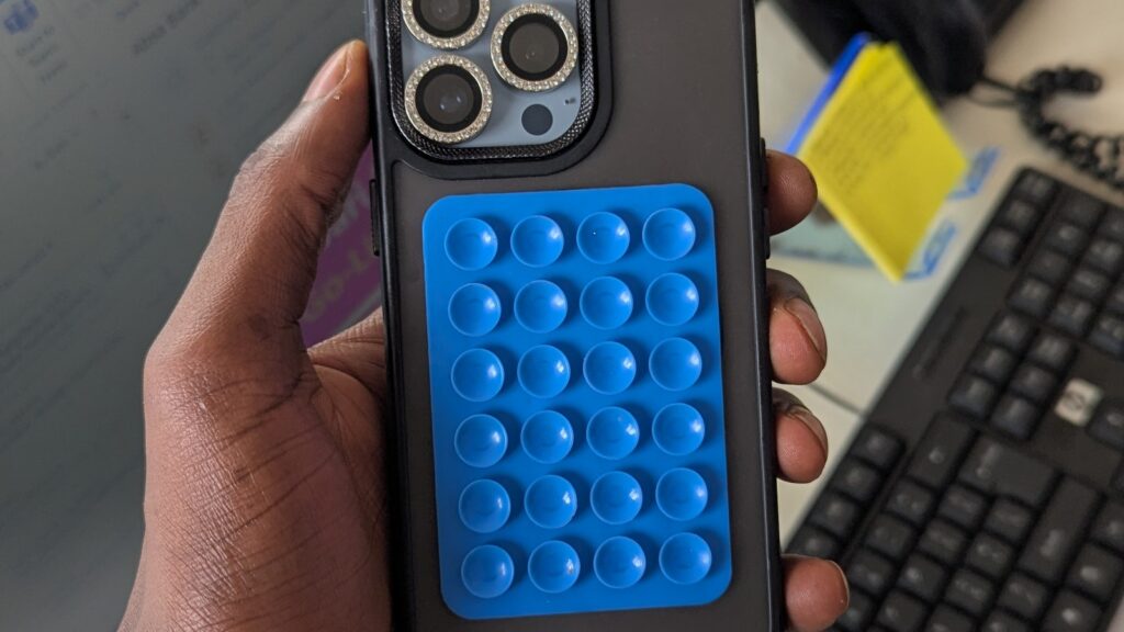 Person holding a smartphone with a blue bubble-popper-style case, in an office with a monitor, keyboard, and paperwork in the background.