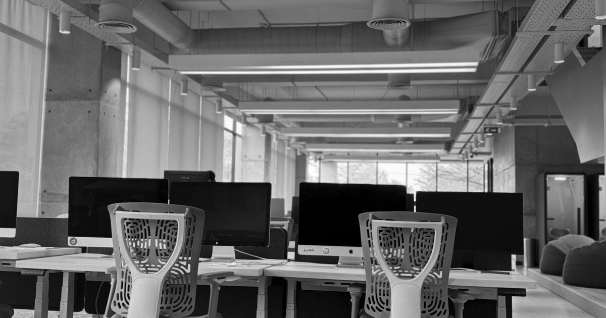 A modern office space featuring several white desks with computer monitors and ergonomic chairs.