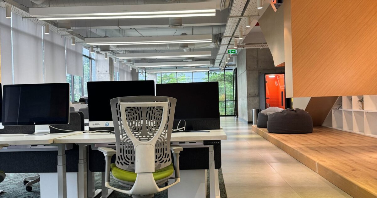 Modern open office with empty desks, ergonomic chairs, large monitors, and natural light from floor-to-ceiling windows.