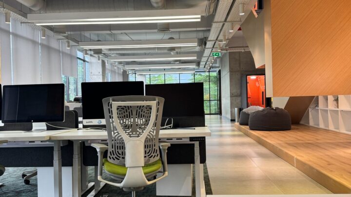 Modern open office with empty desks, ergonomic chairs, large monitors, and natural light from floor-to-ceiling windows.