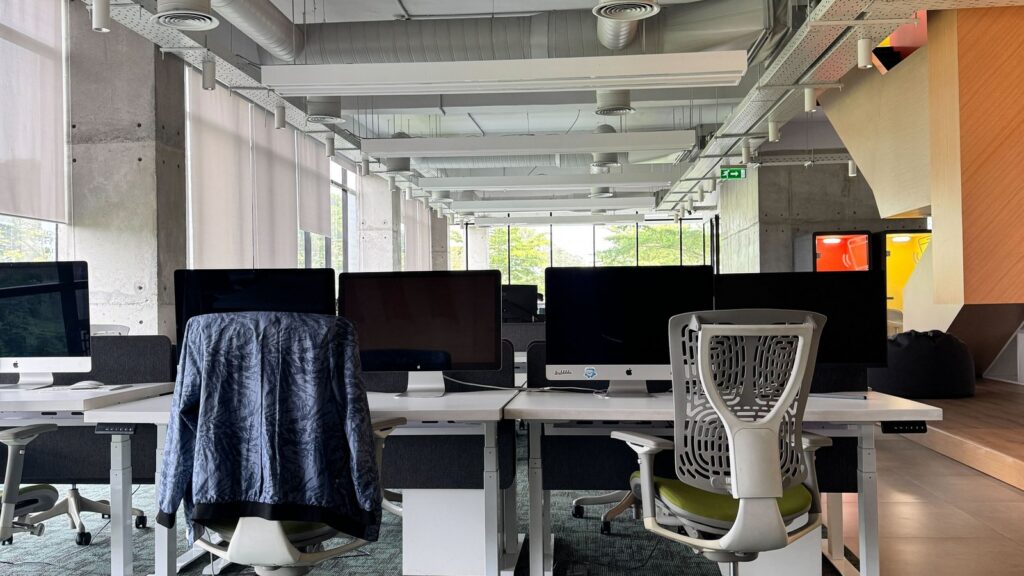 Modern office space featuring rows of desks with computers, ergonomic chairs, and large windows.