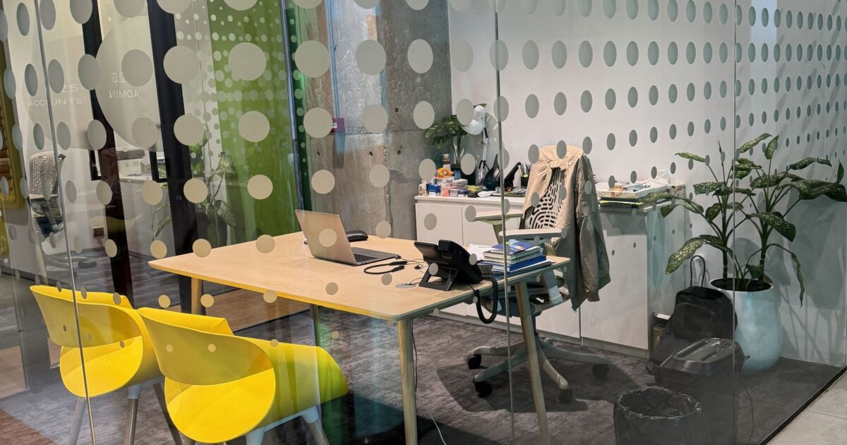 Modern glass-walled office with a wooden desk, open laptop, two bright yellow chairs, and potted plants.