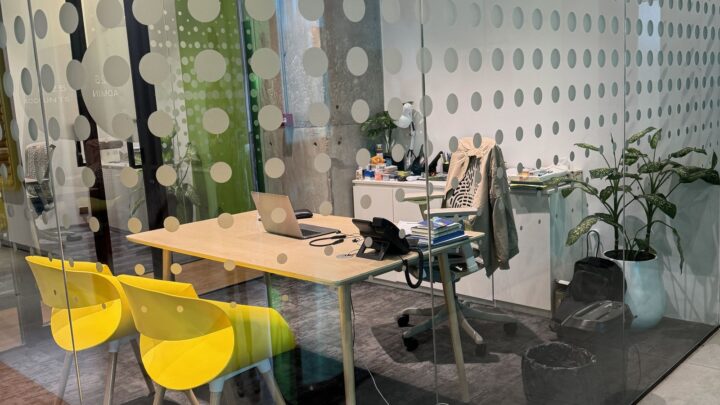 Modern glass-walled office with a wooden desk, open laptop, two bright yellow chairs, and potted plants.